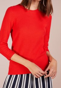Czerwony sweter z dzianiny, okrągłym dekoltem i rękawami o długości trzech ćwiartek. Żebrowany dół i mankiety, teksturowana tkanina. Połączony z paskowanym spódnicą.