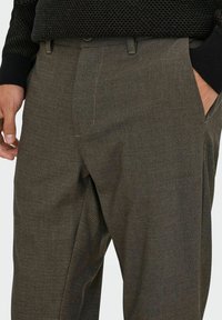 Braune kariertes Hose mit seitlichen Taschen und einem Knopfverschluss. Der Stoff hat eine glatte Textur mit einem dezenten Muster.