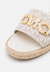 Gros plan d'une sandale blanche à bout ouvert avec semelle en jute tissée et logo MK métallique doré sur une bride en tissu à motifs.