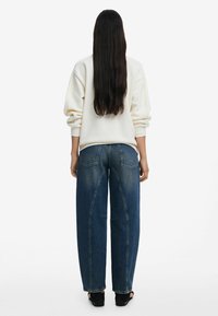 Oversized cream sweatshirt with ribbed cuffs worn over dark blue wide-leg jeans featuring curved seam detailing and a relaxed fit.