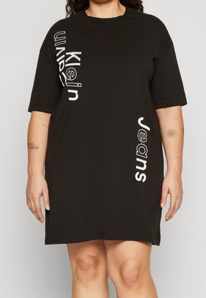 Musta T-paitamekko, valmistettu puuvillasta, jossa on löysä istuvuus, pyöreä pääntie ja pystysuora valkoinen teksti "Klein Jeans" edessä.