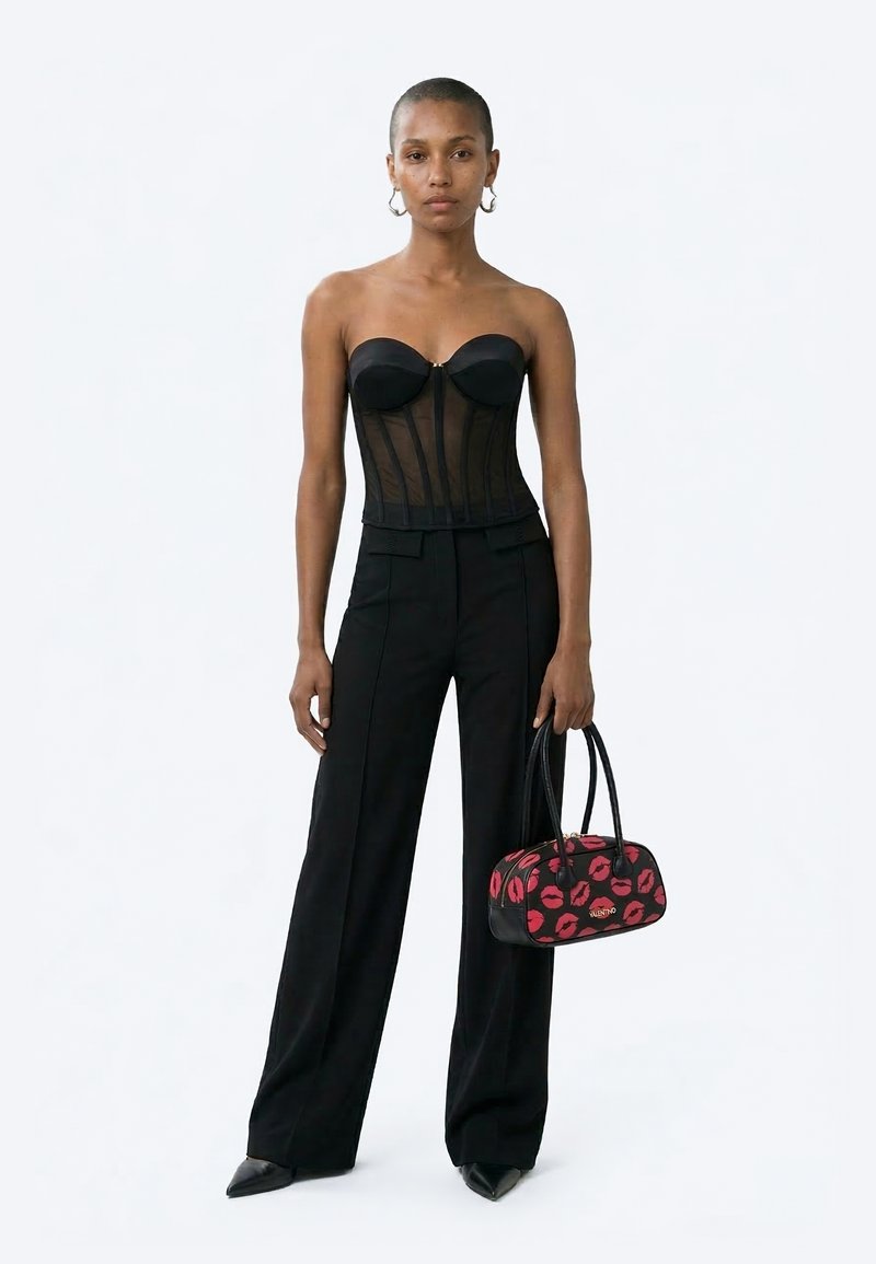 Γυναίκα με μαύρο κορσέ χωρίς τιράντες και παντελόνι με φαρδύ πόδι, κρατώντας μια μαύρη τσάντα χειρός με κόκκινο αποτύπωμα χείλους, στάζει μπροστά σε ένα απλό φόντο.