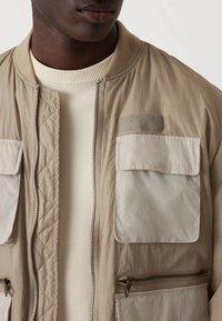 Veste bomber beige avec un design matelassé, dotée de deux poches poitrine, d'une fermeture éclair et d'un patch logo "GUESS" sur l'épaule gauche.