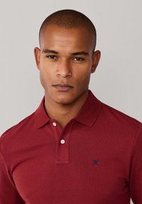 Polo bordeaux en tissu texturé, avec deux boutons au col et un logo bleu marine sur la poitrine. Gros plan sur le visage d'un modèle.