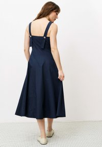 Navyblaues Midikleid mit breiten Trägern, Knopfdetails im Rücken und ausgestelltem Rock. Glattes Gewebe und einfarbig, kombiniert mit beigen Schuhen.