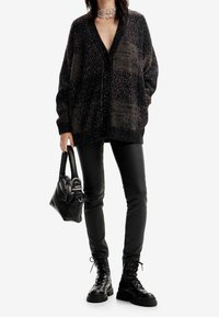 Svart, texturerad stickad cardigan med v-ringning och knappar, matchad med svarta tajta byxor och snörda kängor. Svart handväska i handen.