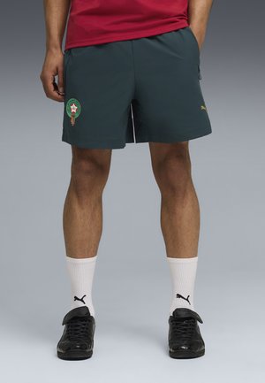 Moški, oblečen v temno zelene kratke hlače s športnim logotipom, bele nogavice Puma in črne športne copate Puma, stoji z rokami v žepih.