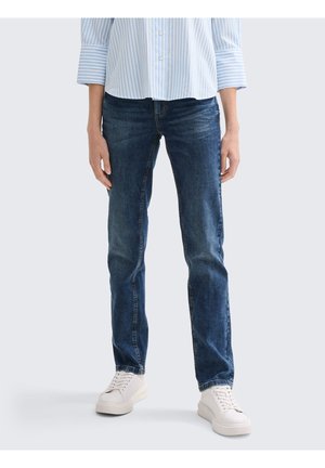 Personne portant une chemise rayée bleue, un jean droit bleu foncé et des baskets blanches à lacets, debout devant un fond clair.