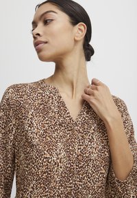 Hnědá a béžová košile s leopardím potiskem, s výstřihem do V, zapínáním na knoflíky a dlouhými rukávy se nařasenými manžetami. Hladká textura látky.