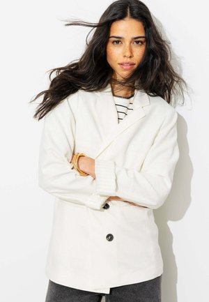 Femme aux longs cheveux foncés portant un blazer blanc, une chemise rayée, un bracelet doré et un pantalon foncé, debout les bras croisés sur un fond blanc.