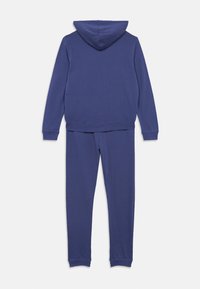 Lillasest kapuutsiga ja sobivate dressipükstega komplekt, mis on valmistatud pehmest kangast. Omab pikki varrukaid, reguleeritavat kapuutsi ja soonikuid ääriseid.