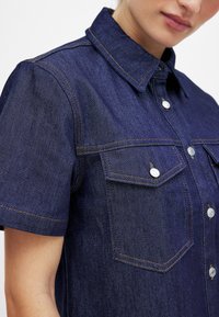 Tume sinine denimist särk lühikeste varrukatega, ees taskud ja kontrastsed oranžid õmblused. Omab kraed ja metallist nuppudega kinnistusi.