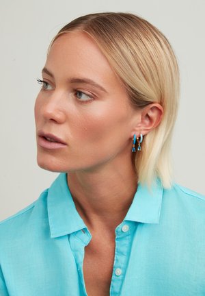 Gold hoop earrings feature a blue accent and triangular drop detail. The model wears a light blue collared shirt with a textured fabric.