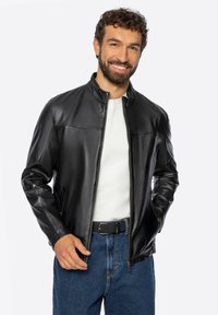 Man die glimlacht en een zwarte leren jas draagt over een wit shirt en een blauwe spijkerbroek, met handen in de jaszakken, tegen een effen lichte achtergrond.