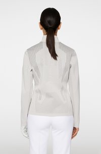 Haut à manches longues gris clair avec des rayures texturées et des trous de ventilation, présentant un col montant et un logo dans le dos, associé à un pantalon blanc.