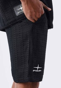 Pantaloni corti neri con un motivo tessuto texturizzato, dotati di una tasca laterale e un logo ricamato bianco. L'orlo presenta un dettaglio sfilacciato.