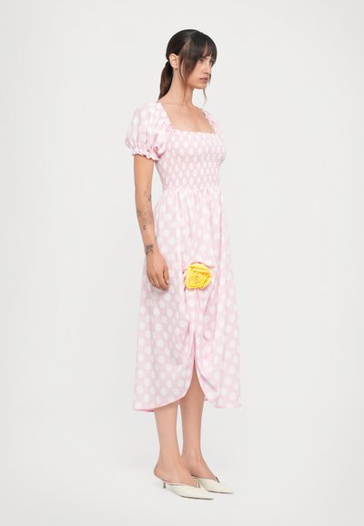 Lyserød kjole med prikker, pufærmer, smocked bodice og en gul stofrose detalje ved hoften. Kjolen falder under knæet.