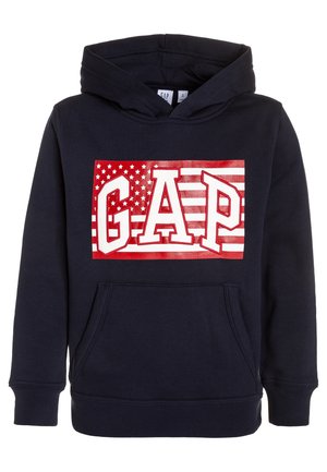 Sweat à capuche bleu marine avec poche avant et motif rouge et blanc comprenant le texte "GAP" sur un fond stylisé de drapeau américain.