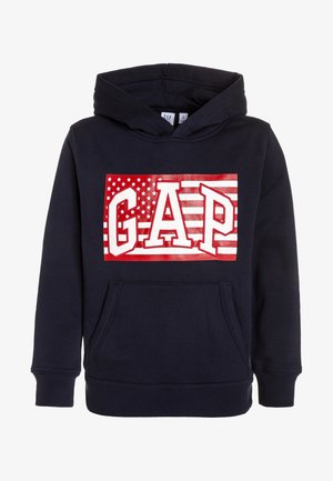 Sweat à capuche bleu marine avec poche avant et motif rouge et blanc comprenant le texte "GAP" sur un fond stylisé de drapeau américain.