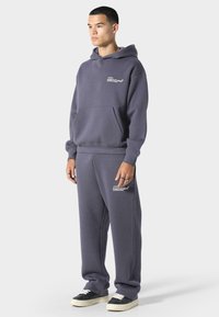 Šedá mikina s kapucí a přední kapsou, se bílým logem, doplněná o šedé tepláky se stejným logem, nošená s černobílými teniskami.