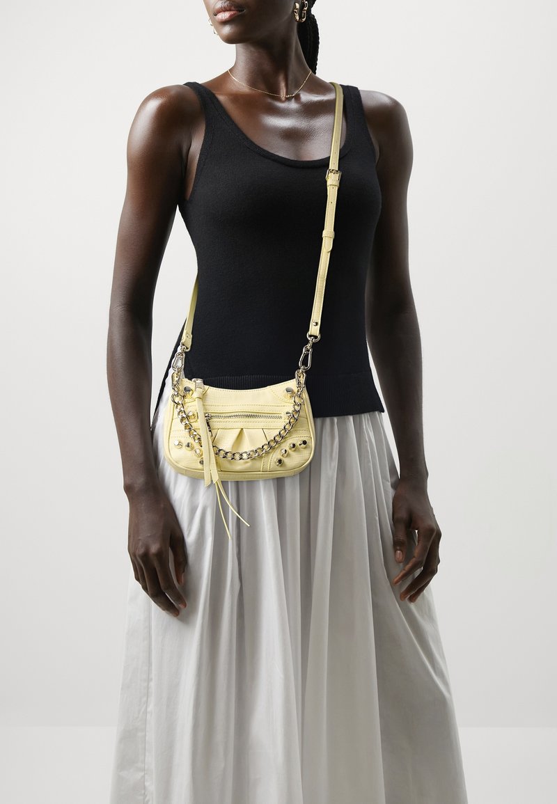 Femme portant un haut noir sans manches et une jupe blanche avec un petit sac bandoulière jaune orné de chaînes et de clous.