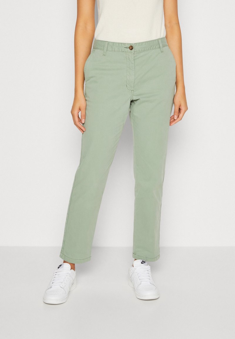 Personne portant un pantalon droit vert clair et des baskets blanches, debout devant un fond clair uni.