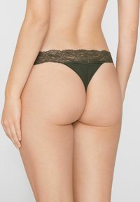 Olivgrüner String mit einem Spitzenbund, der eine glatte Textur und minimalen Stoffanteil bietet. Das Design verfügt über einen hochgeschnittenen Rücken und dünne Träger.