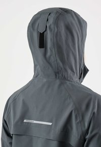 Persona che indossa una giacca impermeabile con cappuccio di colore grigio scuro con logo riflettente "Swedemount" sul retro, vista da dietro su sfondo bianco.