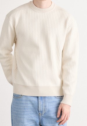 Person, die einen cremefarbenen gerippten Strickpullover und hellblaue Jeans trägt und vor einem schlichten hellen Hintergrund steht.