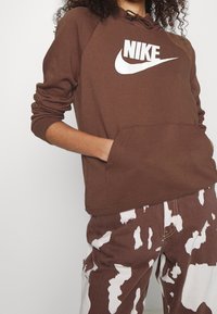 Brown Nike sweatshirt with a white logo, long sleeves, and a hood. Paired with white and brown patterned pants. Front pocket detail visible.