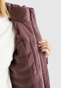 Brun isolerad jacka med en kviltad yttersida och ett mjukt fluffigt innerfoder; har en sidoficka med dragkedja.