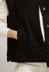 Gilet en laine noire présentant une coupe décontractée, une base arrondie, de grandes poches avant et une fermeture à boutons. Tissu texturé avec une apparence douce.