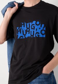 Persona vestida con una camiseta negra con texto abstracto azul, sosteniendo una chaqueta de mezclilla sobre un hombro y con una mano en el bolsillo del pantalón.