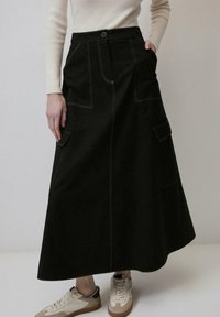 Jupe midi noire avec des détails de couture visibles, deux poches avant et une taille ajustée, associée à des baskets légères. Texture en denim lisse.