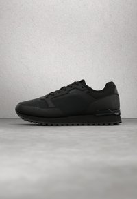 Baskets basses noires avec un design texturé, fermeture à lacets et logo "BOSS" sur le côté, présentées sur une surface grise et un fond uni.