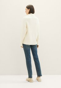 Suéter de punto acanalado crema con cuello alto, combinado con jeans de mezclilla azules y zapatos de plataforma beige, visto desde la parte de atrás.