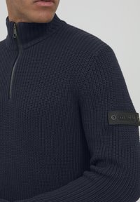 Maglione lavorato a maglia blu navy con colletto alto, mezza cerniera e tessuto strutturato. Presenta una toppa nera sulla manica sinistra con testo stampato.