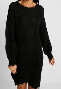 Abito lavorato a maglia nera con una vestibilità ampia, maniche lunghe e bordi sfrangiati. Tessuto testurizzato, collo tondo, lunghezza al ginocchio, caratterizzato da un design a punto aperto.