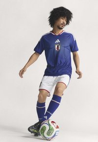 Blå fotbollströja med texturerat mönster, vita shorts med röda detaljer, blå strumpor och svarta fotbollsskor, med ett emblem för Japan och en mönstrad fotboll.