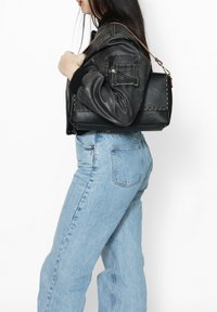 Zwarte leren handtas met een textuur oppervlak en zichtbare stiksels, gedragen op één schouder; gestyled met een zwarte bomberjack en een blauwe jeans.