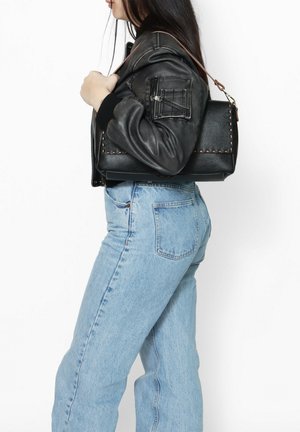 Sac en cuir noir avec une surface texturée et des détails de couture visibles, porté sur une épaule ; stylé avec une veste bomber noire et un jean bleu.