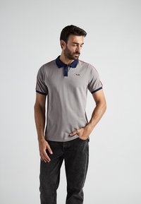 Szara koszulka polo z granatowym kołnierzykiem i czerwonymi paskami na ramionach. Krótkie rękawy i małe logo na lewej piersi. Noszona z ciemnymi dżinsami.