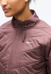 Κοντινή εικόνα ενός ατόμου που φοράει ξυρισμένο μπουφάν μάβης με ψηλό γιακά και λευκό λογότυπο της Nike στο στήθος.