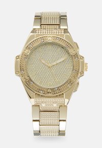 Gullur klokke med rund urskive, prydet med steiner, teksturert gullring og mønstret bezel. Klokken har luminerende visere og timemarkører.