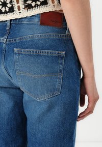 Vaqueros de mezclilla azul con una textura resistente, que presentan un parche de cuero marrón con el logo y detalles en el bolsillo trasero. Diseño suave y ajustado.