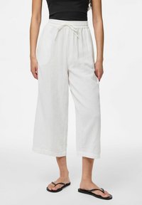 Pantalons blancs à jambe large fabriqués en tissu texturé, dotés d'une taille élastique avec cordon de serrage et de deux poches latérales. Tongs visibles.