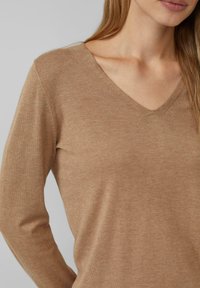 Kobieta ubrana w beżowy sweter z dekoltem w serek, długi rękaw, skrócony tak, aby odsłonić ramiona i górną część torsu na jednolitym tle.