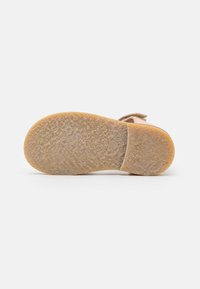 Sula av skor gjord av texturerat gummi i beige färg. Har en slät, rundad form och ett logotryck i mitten.