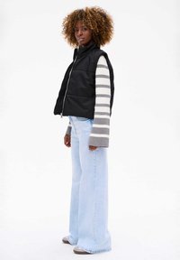 Czarna kamizelka puchowa z wysokim kołnierzem i przednim zamkiem, noszona na szarej i białej bluzce z długim rękawem w paski. Jasnoniebieskie dżinsy w fasonie dzwonów.