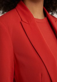 Červený přiléhavý blazer s hladkým materiálem a ostrými klopami, vrstvený přes strukturovanou červenou košili. Žádné viditelné kovové prvky ani vzory.